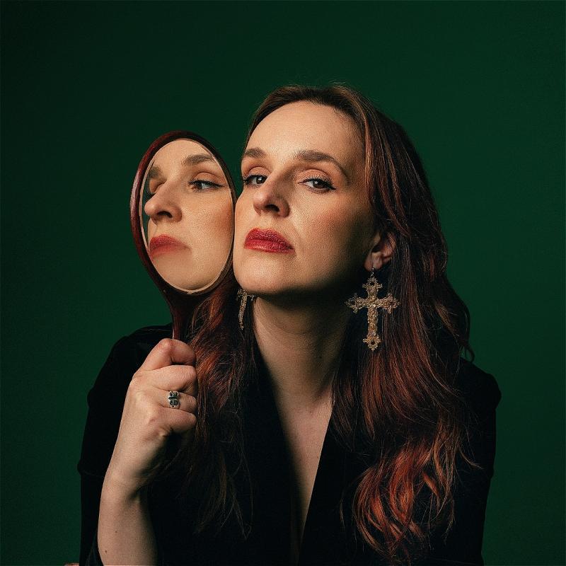 Katie Norris, a white woman with long brown hair, wearing red lipstick, gilded crucifix earrings and a black top, is looking at the camera holding a hand mirror to the side of her face so that the reflection of her face is looking to the side.