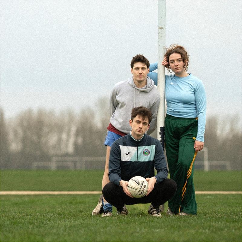 Three individuals stand on a muddy football pitch. One is in front holding a football and squatting, while the other two are leaning against a goalpost.