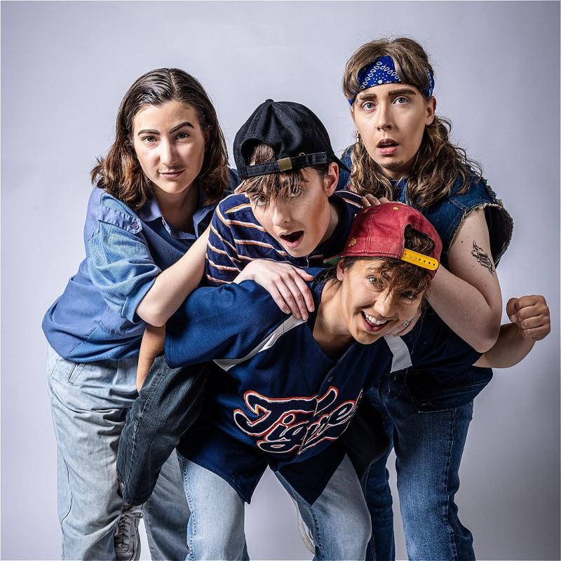 Four drag kings wearing 90s clothing stand huddled together posing like a stereotypical boyband. They look cheeky and youthful.  
