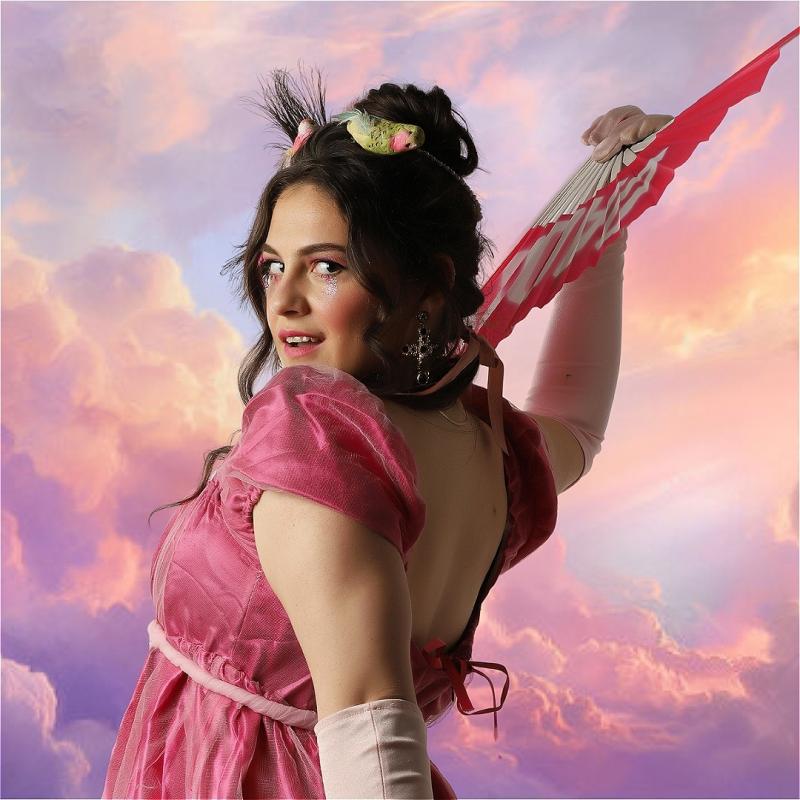 Rosalie Minnitt, a white woman with long dark hair is looking over her shoulder at the camera, smiling. She is wearing a pink princess dress and long white gloves, as well as a red fan. She has fake birds in her hair and glittery pink makeup around her eyes. Behind her is a beautiful pink and blue cloudy landscape. 