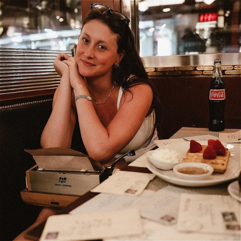 A woman sat in a warmly lit restaurant smiles at the camera. She is sat on a table with a typewriter on her lap. On the table there is a plate of food, a glass bottle of coca cola and a collection of letters.