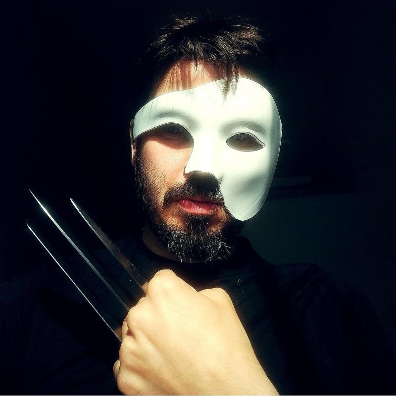 Joz Norris, a white man with brown hair and beard, is looking at the camera wearing a "phantom of the opera" half-face white mask. He is holding his left hand up on his chest with three knives between his fingers, looking like Wolverine's hand.