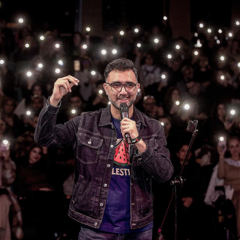 The performer holds a microphone in his left hand with an audience behind him holding their phone flashlights up