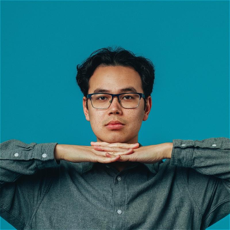 A man wearing glasses in a long sleeve shirt stares at you with his hands interlocked under his chin. 