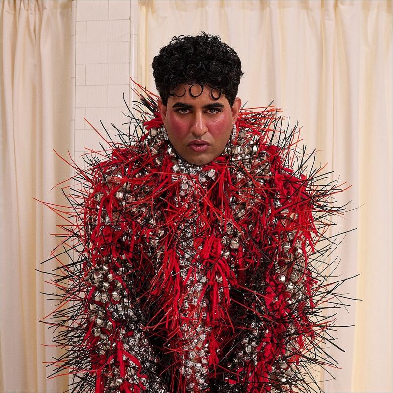 ALOK, a non-binary brown person with short curly black hair and blush on their cheeks, is looking directly at the camera. They are wearing a fashionable coat made of red and black spikes and silver jingle bells, covering their body, neck and hands. The background is made of cream colour curtains.