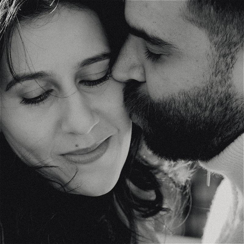 A close-up picture of a brown man with a beard and a brown woman with long black hair. The woman is facing the camera but looking down and smiling as the man is kissing her cheek, eyes closed.