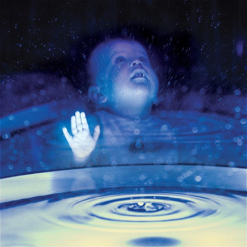 A baby presses their hand against a glass surface, looking up in wonder. In the foreground a pool of water seems to be forming.