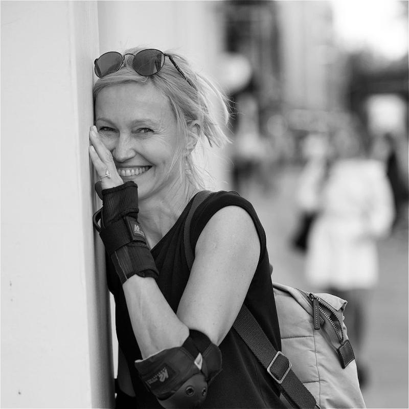 A black and white photo of Ingeborga Dapkunaite, a white woman with blonde hair, sunglasses on her head, a sleeveless black top and wrist and elbow braces. She is looking at the camera smiling, her left hand and side of her face resting on a wall. The background is a blurred street with silhouettes and buildings.
