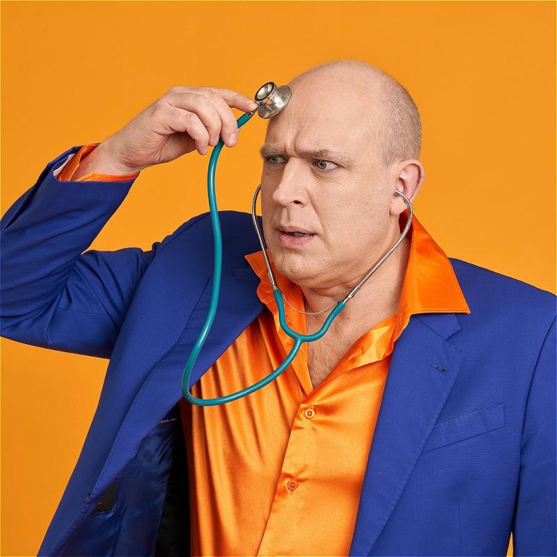 A bald Man dressed in a bright blue and orange suit holds stethoscope to his head. He pulls a concerned expression as he listens to the contents of his brain.