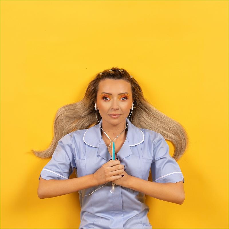 A woman with long hair in a nurses outfit lies on the floor with headphones in. 