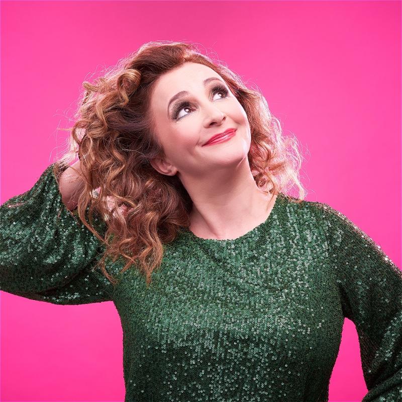 Lucy porter stands in a sequin green long sleeve top, with curly blonde hair looking into the top corner on a pink background. 