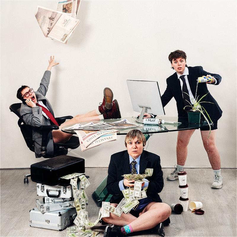 Three people in suit jackets and no trousers sit at a glass table. One shouts loudly on the phone. Another is on a computer whilst watering a plant with an energy drink. The other sits on the floor with loads of money in their hands. 