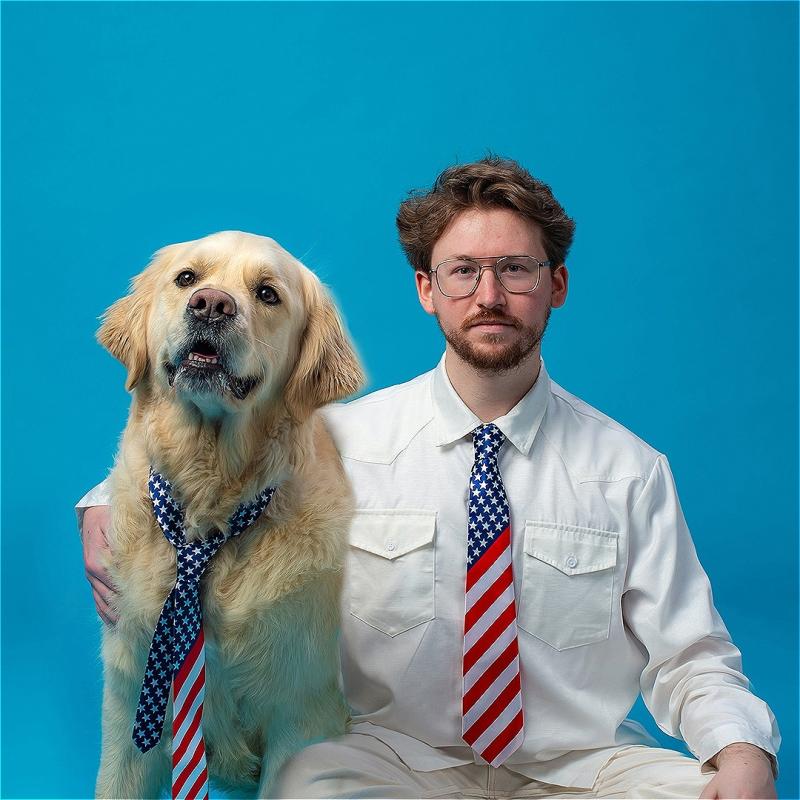 A man and his golden retrierver sit in front of a bright blue background. He wears a shirt and an American flag tie. His dog also wears an american flag tie.  