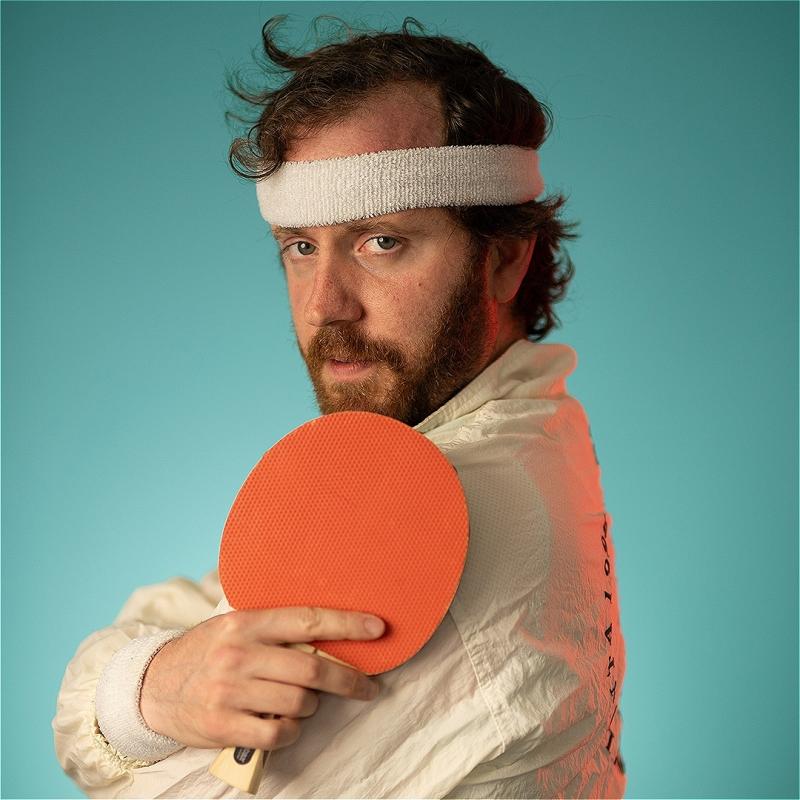 A man in a white shell jacket, holding an orange ping pong bat and wearing a white headband looks at us with intrigue. 