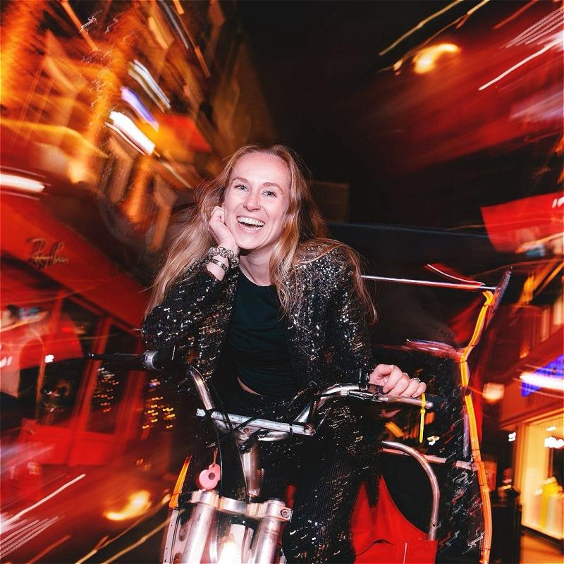 A woman with long blonde hair rides through the streets of London at night on a tuk tuk. 