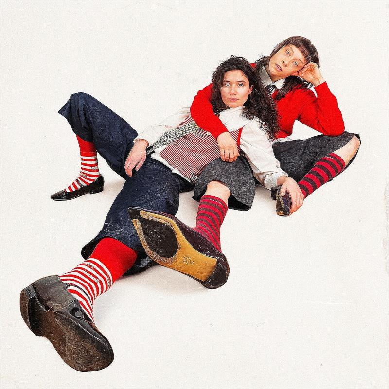 Two female presenting people sit on the floor in a relaxed manner. One lies across the other. They are wearing stripey socks, blue/black jorts, white shirts and ties. It looks very preppy. 