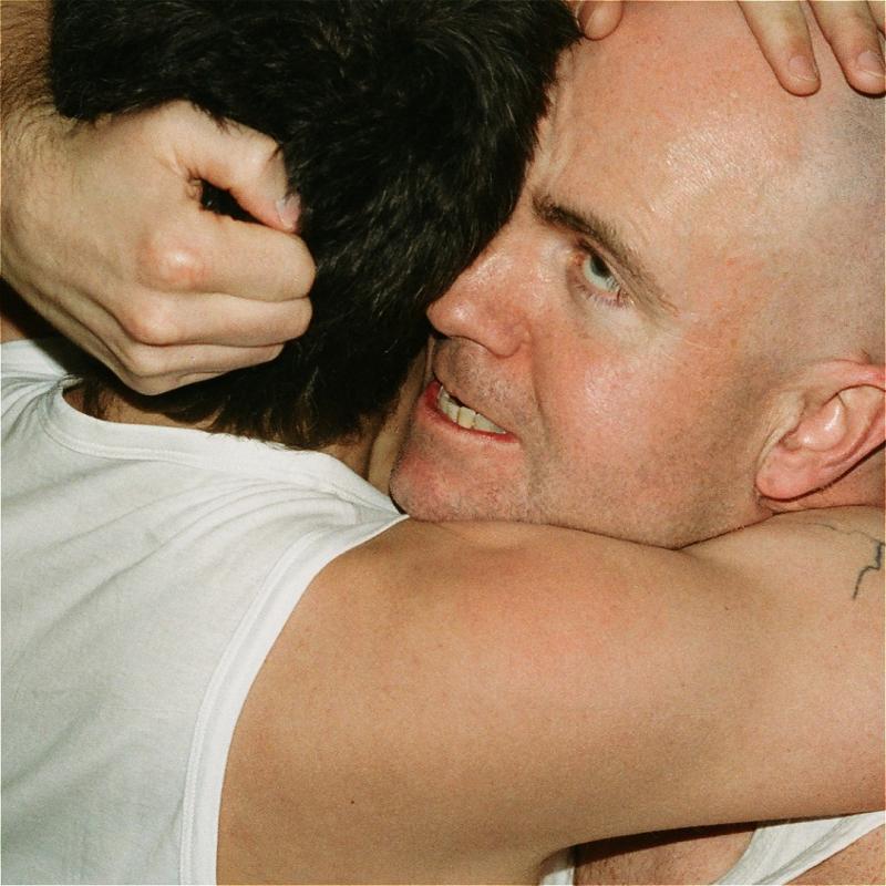 A caucasian bald man and a man with dark hair in white vests embrace with a look of possession