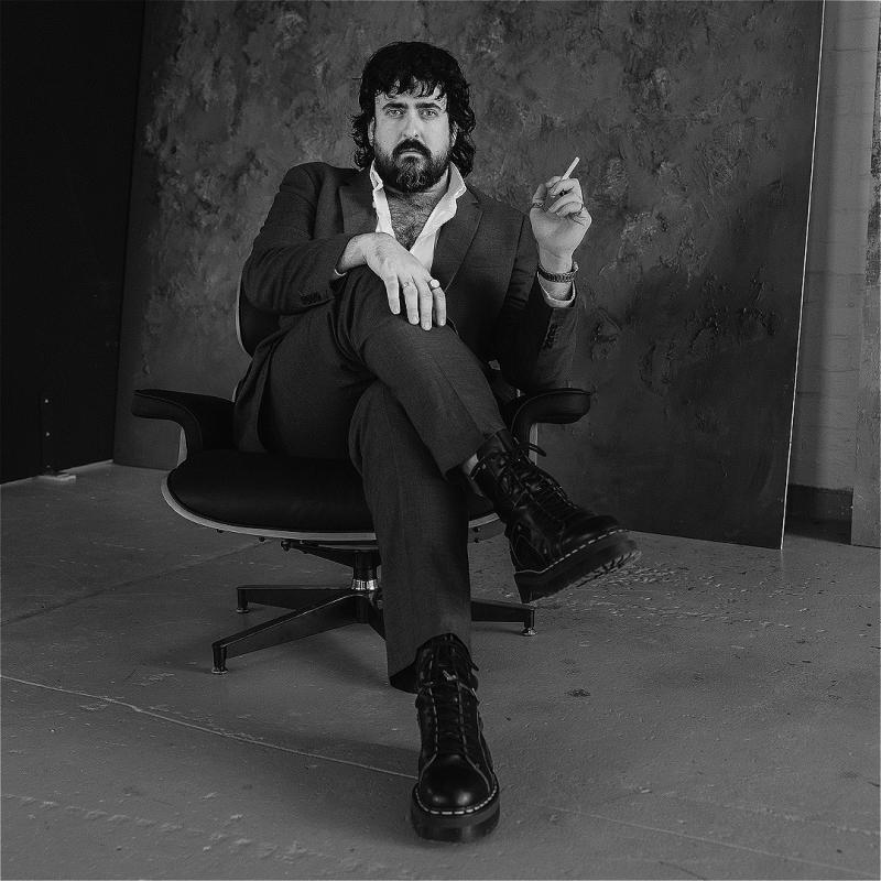 A black and white photo of Zachary Ruane, a white man with a black mullet and beard, wearing a grey suit, white shirt and chunky black boots. He is seriously looking at the camera, holding a cigarette in his left hand and sitting in an office chair. 