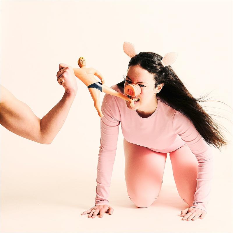 A woman dressed as a pig holds a ken doll in her mouth, whilst a man flexing his arm holds the other side of the doll. 
