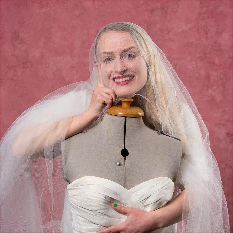 A woman stands behind a mannequin with a wedding dress on it. She has a veil on her head, and looks manically at you.