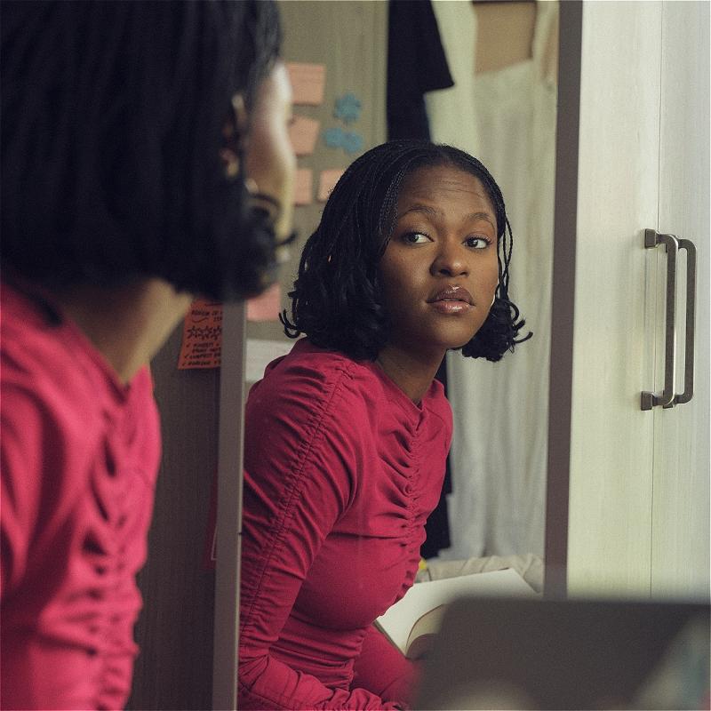 Ayoade Bamgboye, a black woman with shoulder length locks and a red long-sleeve top, is looking at herself in the mirror of a bedroom wardrobe. In the background we can see post-it notes, a white dress and her laptop.