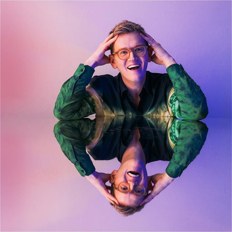 Chris Parker, a white man with round glasses and short blond hair, is smiling at the camera with his hands in his hair. He is resting his elbows on a mirrored surface, making the lower half of the photo his upside-down reflection.