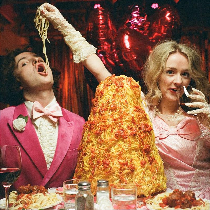 Two individuals dressed in pink sit at a table with a giant pile of spaghetti on it. One of the individuals is enthusiastically feeding a piece of the spaghetti to the person sat next to them whilst sipping a glass of red wine. In the background balloons in the shape of the number 18 can be seen.