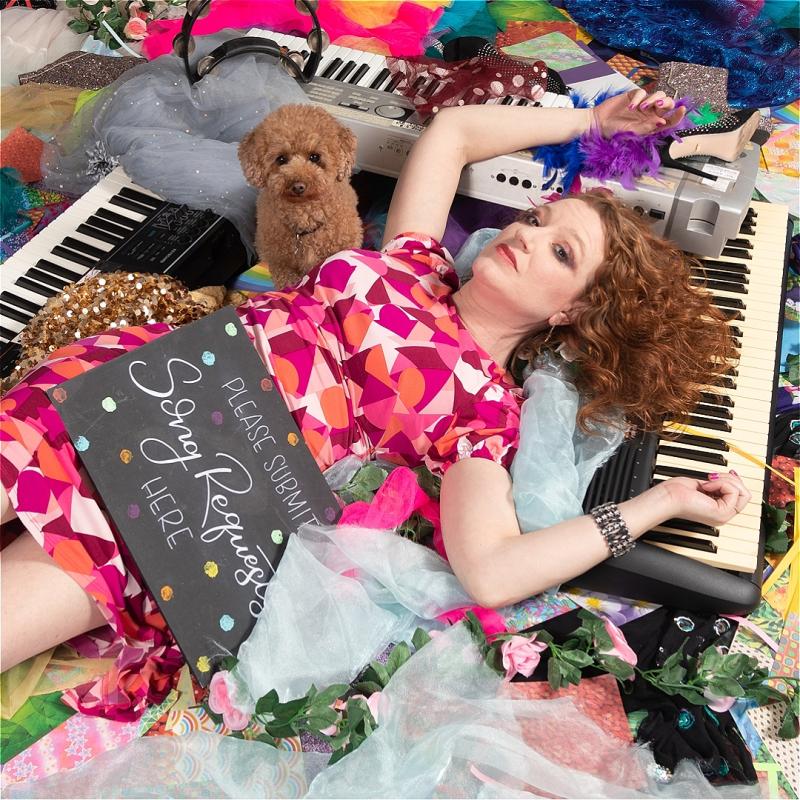 An individual lies amongst a pile of roses, keyboards and sequins. A chalk board rests against her lap with the words 'Please Submit Song Requests Here' written on it.