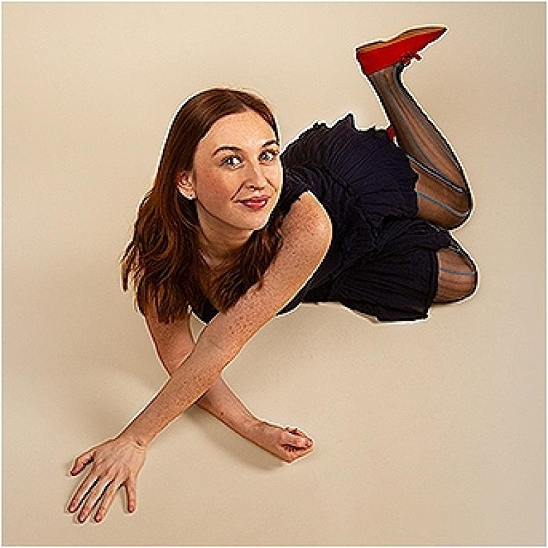 A woman wearing a black dress and red shoes lies across the floor. She is smiling warmly at the camera and has one leg pointing up.