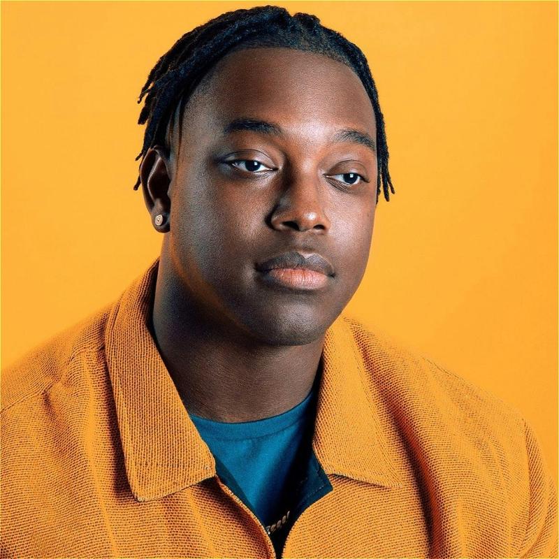 Ify, wearing an orange shirt against an orange background, looks blankly off. 
