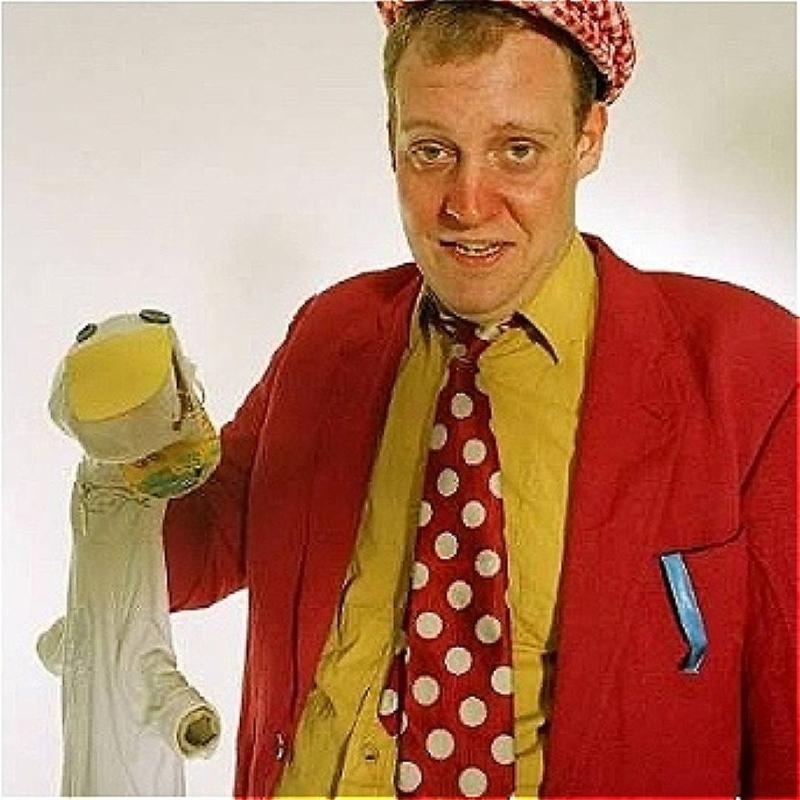 Jeremy Lion, a white man with short ginger hair, wearing a red flat cap, a yellow shirt with a red jacket and a red polka dot tie, is looking at the camera holding a white sock puppet over a white background.