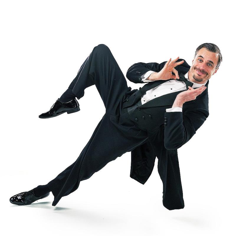 A man in a black morning suit and bow tie, with slicked back hair, twizzles his bowtie and leans on one foot to the right, at a dangerous angle.
