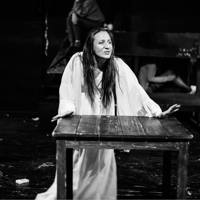 A black and white image of an individual with long black hair mid speech. Their palms are pressed against a wooden table and they are dressed in a long white nightgown.