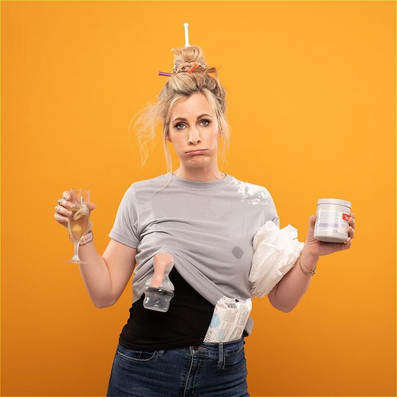 A tired woman stands in front of an orange background. She is holding a pot of lotion in one hand and a glass of champagne with a dummy in it in the other. A packet of wipes can be seen sticking out of her jeans and a breast pump hangs from her dirty T shirt.
