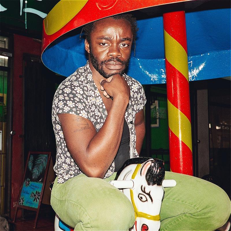 Demi Adejuyigbe, a Black person with short afro hair and a beard, wearing a flowery shirt and green trousers, is sitting on a small carousel horse, looking seriously at the camera. Over him is the umbrella shaped top of the carousel and the background is a shopping centre.