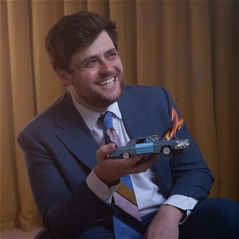 Bróccán Tyzack-Carlin, a man with brown hair and stubble, is wearing a blue suit and white shirt with a colourful tie. He is smiling looking to the side and holding a miniature car with the hood on fire, over a brown curtain background.