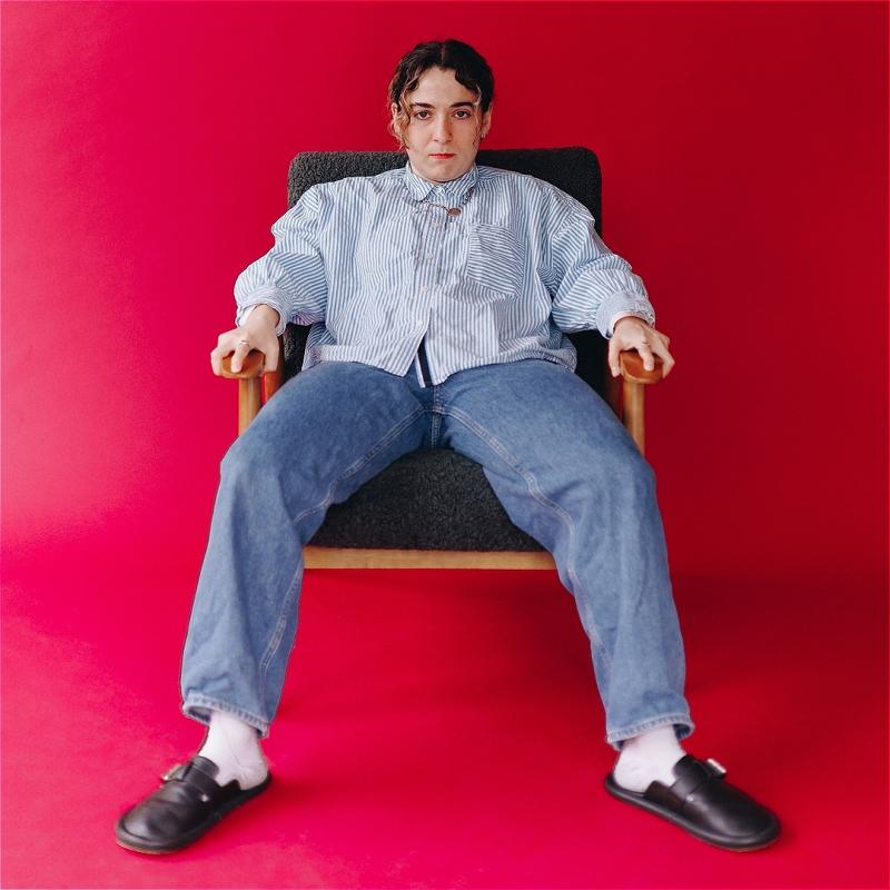 An individual is slumped on a chair and staring expressionless at the camera. The floor and wall are painted a vivid red.