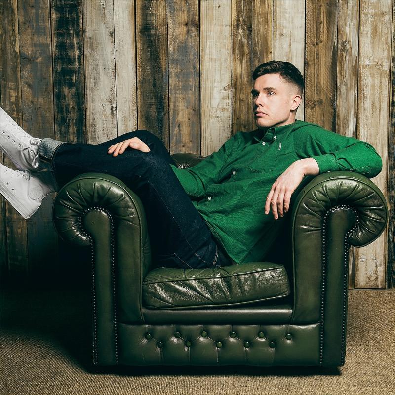 Ed Gamble lounging on a green leather armchair staring into the distance with a forlorn expression on his face.