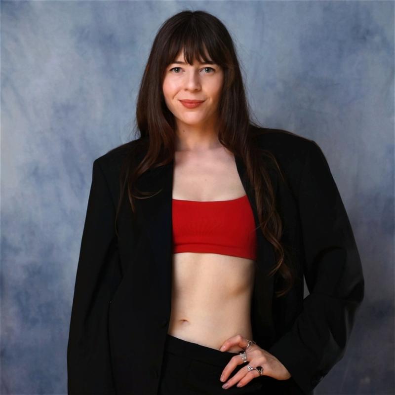 Chelsea, wearing a red sports bra over a black blazer, smiles at the camera before a grey background.