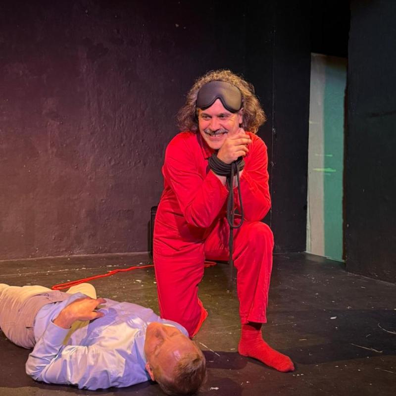 A man wearing an all red outfit crouches next to a body. His hands are bound but he smiles out into the audience.