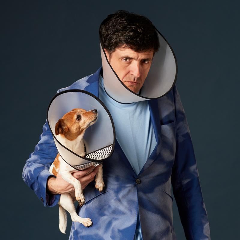 Spencer Jones wears a dog cone and blue suit and holds a small dog also wearing a cone. 