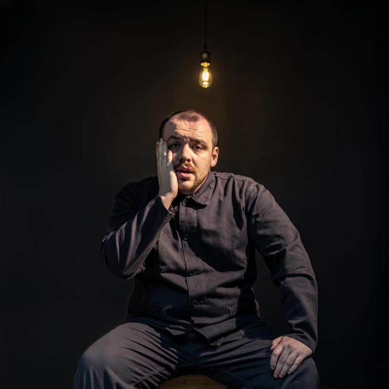 A person is in a dark empty room, beneath a start bare lightbulb. They're wearing all-black and they look worried, holding a hand up to their bearded face. 
