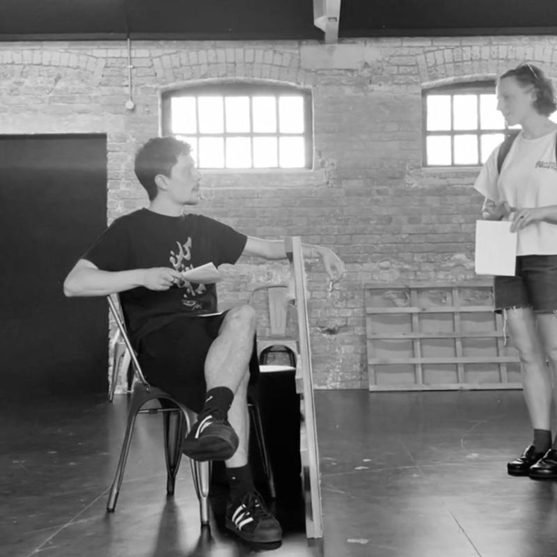 A black and white image of two people seemingly rehearsing. One is sat down while the other is stood. Both of the performers are holding a piece of paper.