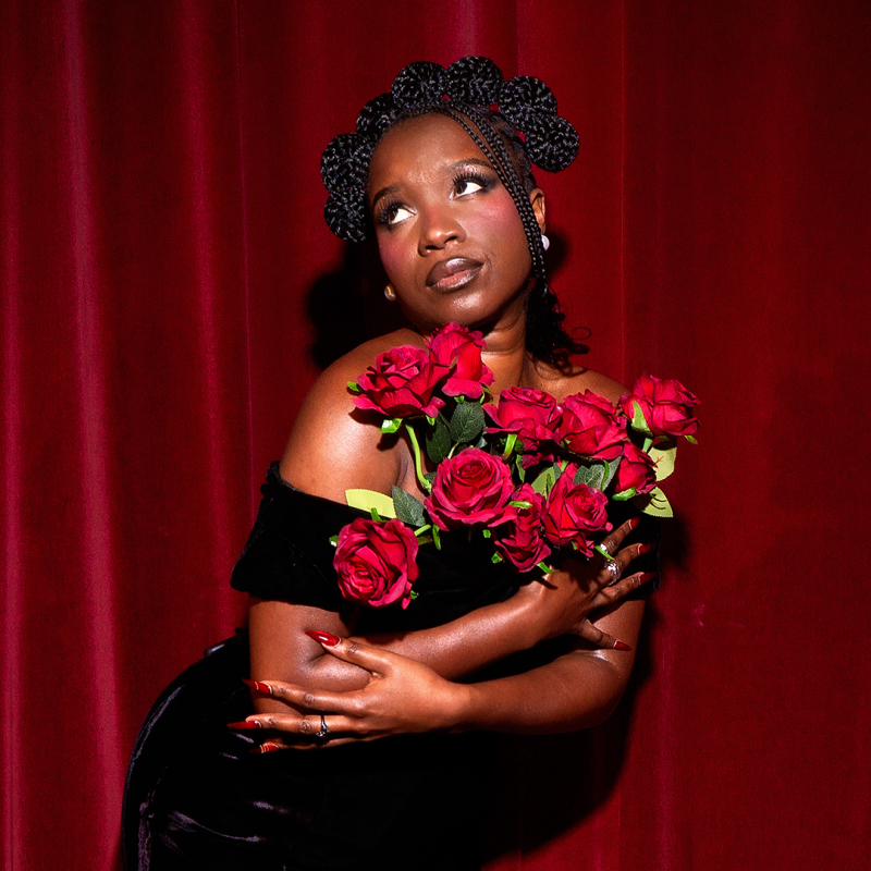 Temi stands against a red curtain, wearing a black dress and holding a large bouquet of red roses