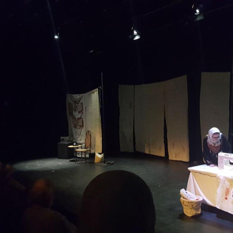 An image of a stage with sheets hung across the back. Centre stage is a table with a sewing machine. A woman dressed in a headscarf is crouched over the table seemingly sewing.