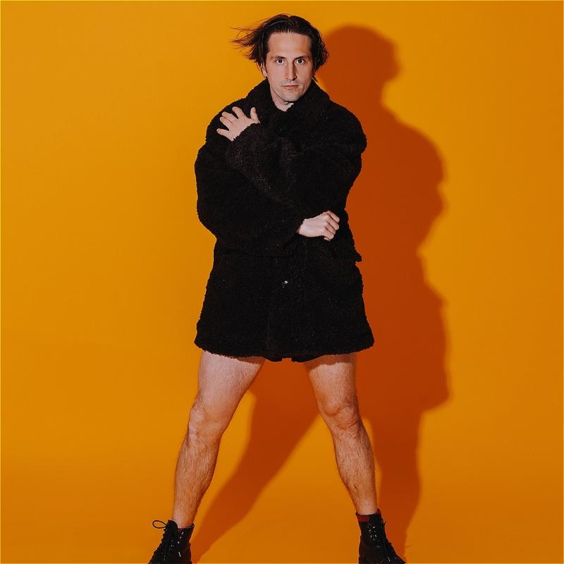 Luke stands against a bright orange backdrop, wearing just a black fluffy coat and some black boots. 