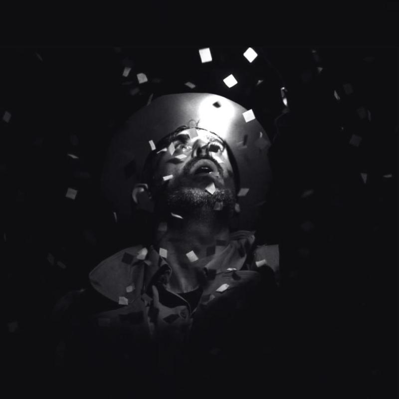 A man in darkness looks up at a bright light from above. Confetti falls around him. 