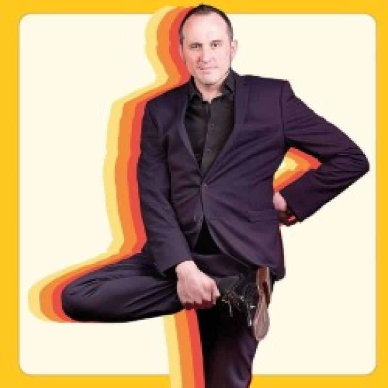 Phil looks into the camera whilst holding up their right foot. Behind them are colourful silhouettes. A