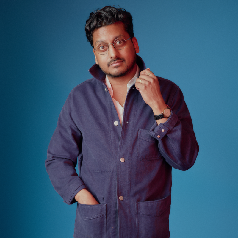 Ahir satnds in front of a bright blue back drop. They are looking calmly into the camera as they fiddle with the collar of their blue boiler suit.