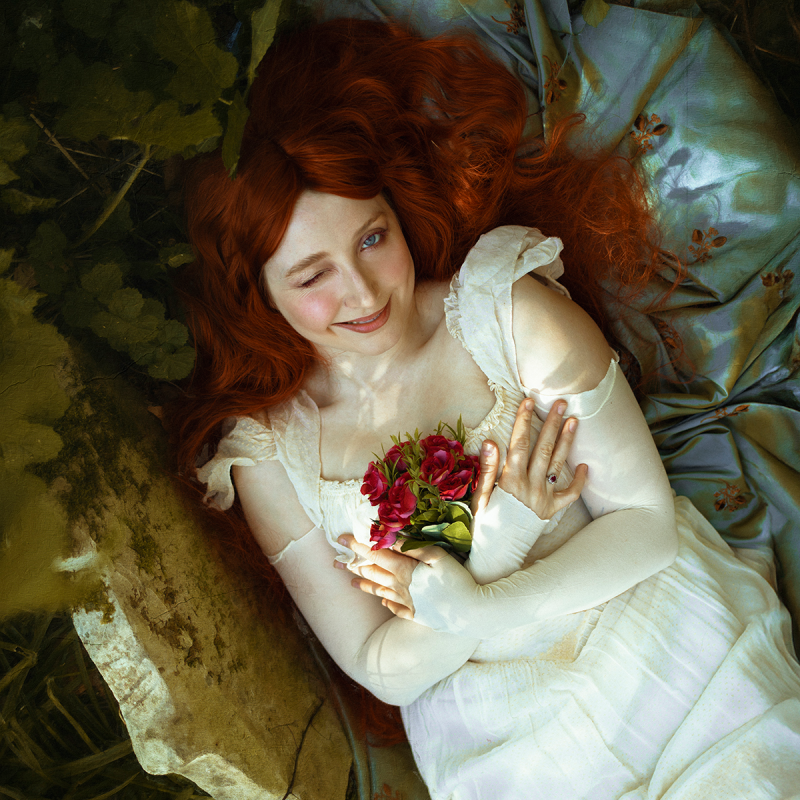 Bebe laying in what appears to be some kind of casket with her red hair flowing against the leaves and holding some red flowers. She may be laying in a grave.